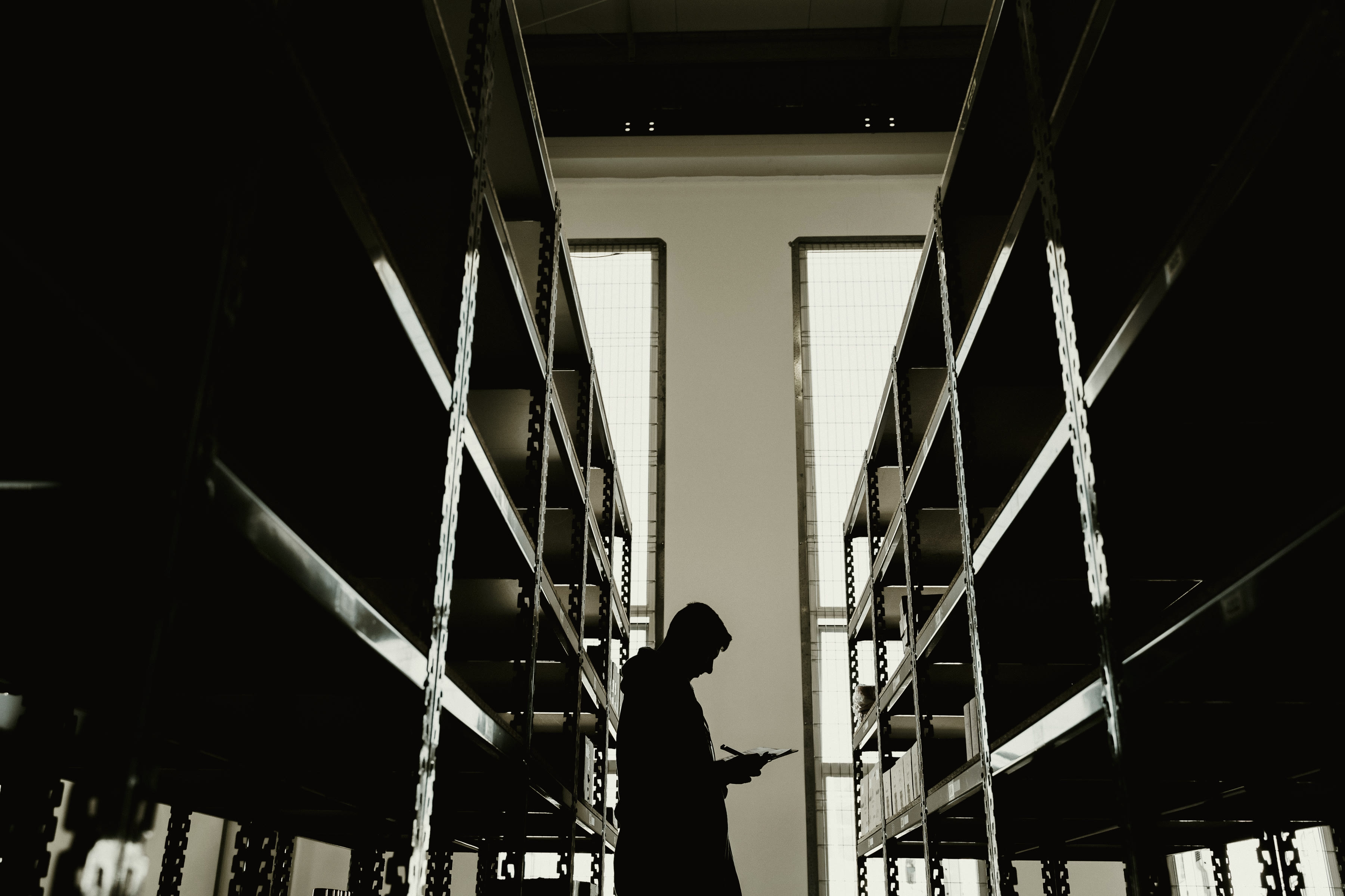 A figure between tall storage racks in a records room, backlit by high windows.